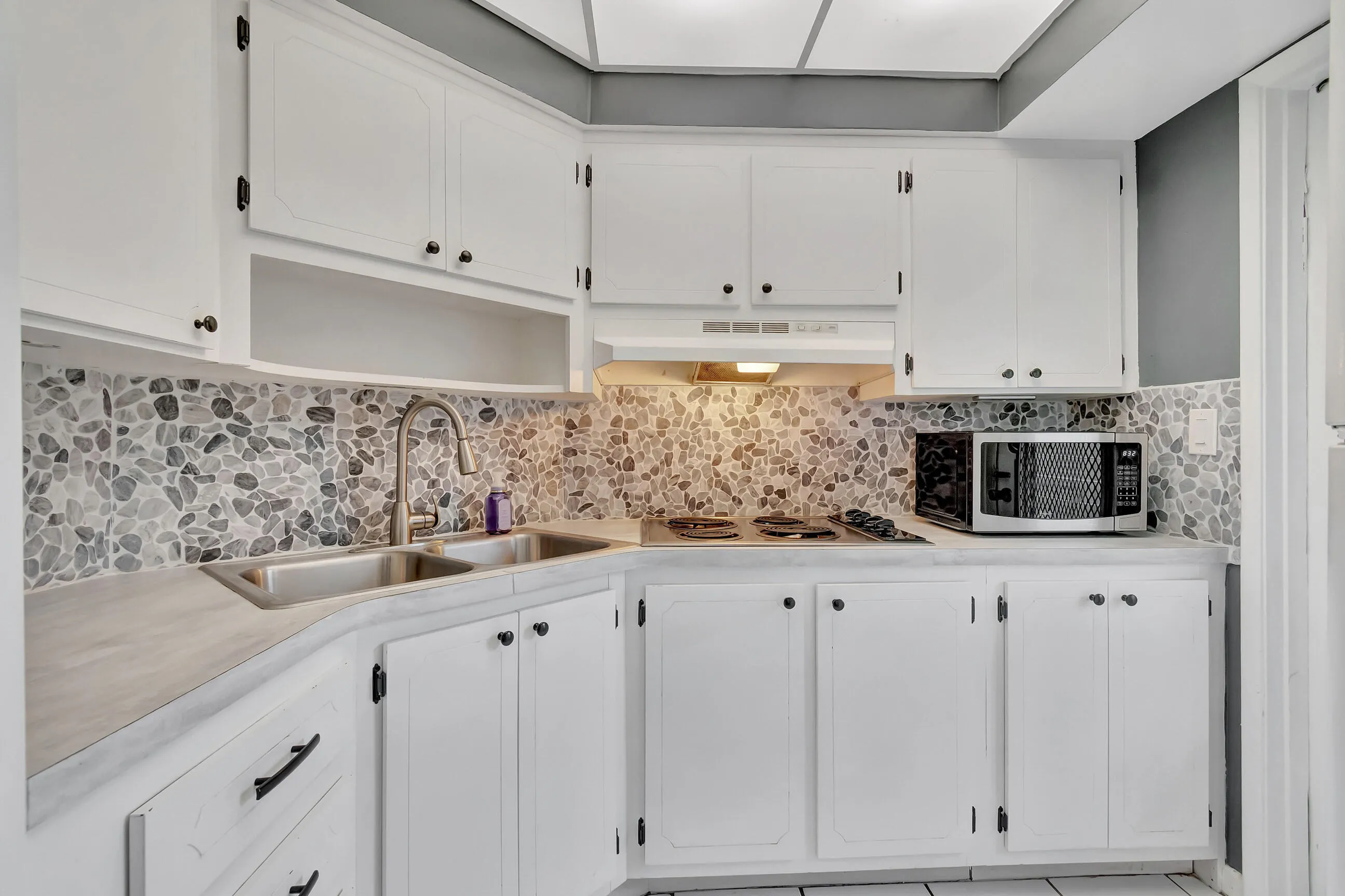 White shaker-style cabinetry with black hardware, stone tile backsplash, stainless steel double basin sink with gooseneck faucet, and light-toned countertops