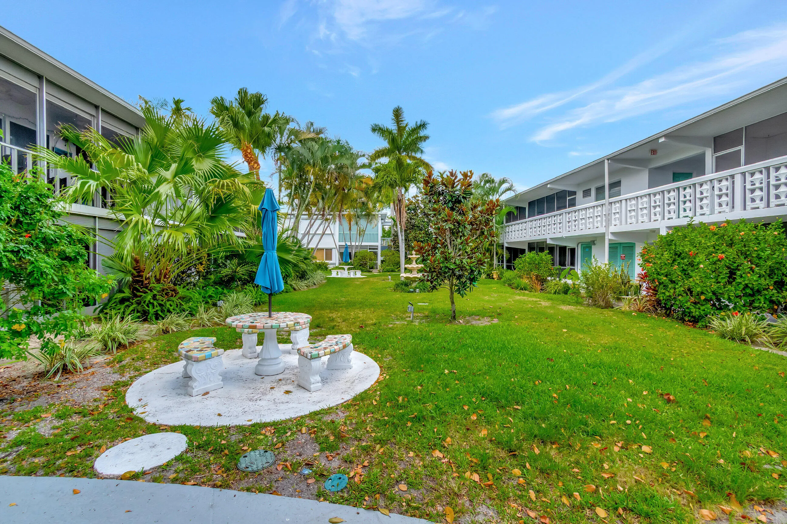 Expansive lawn area featuring a stone patio with seating, mature palm trees, diverse landscaping, and a decorative fountain