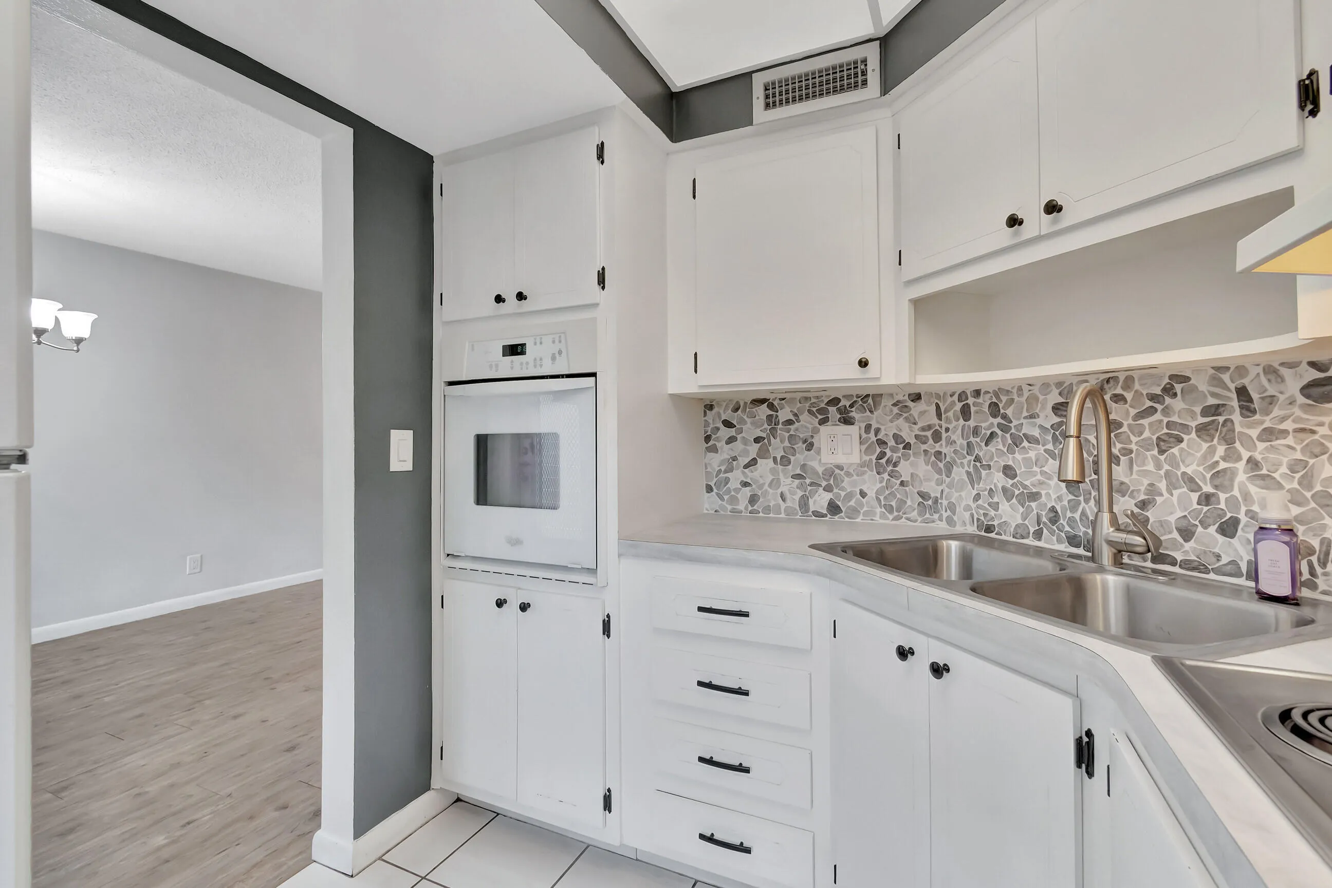 White cabinetry, pebble tile backsplash, dual basin stainless steel sink, built-in oven, and wood-finish flooring