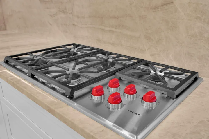 Stainless steel Wolf gas cooktop with red control knobs, set into a stone countertop with a matching stone backsplash, and white cabinetry