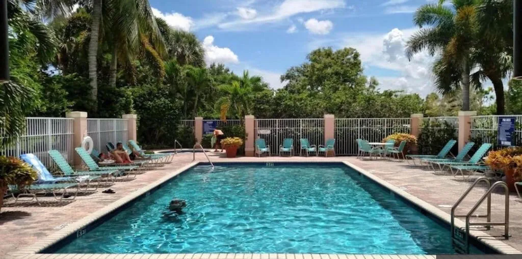 Rectangular swimming pool with a surrounding paver deck, white perimeter fencing, and mature tropical landscaping