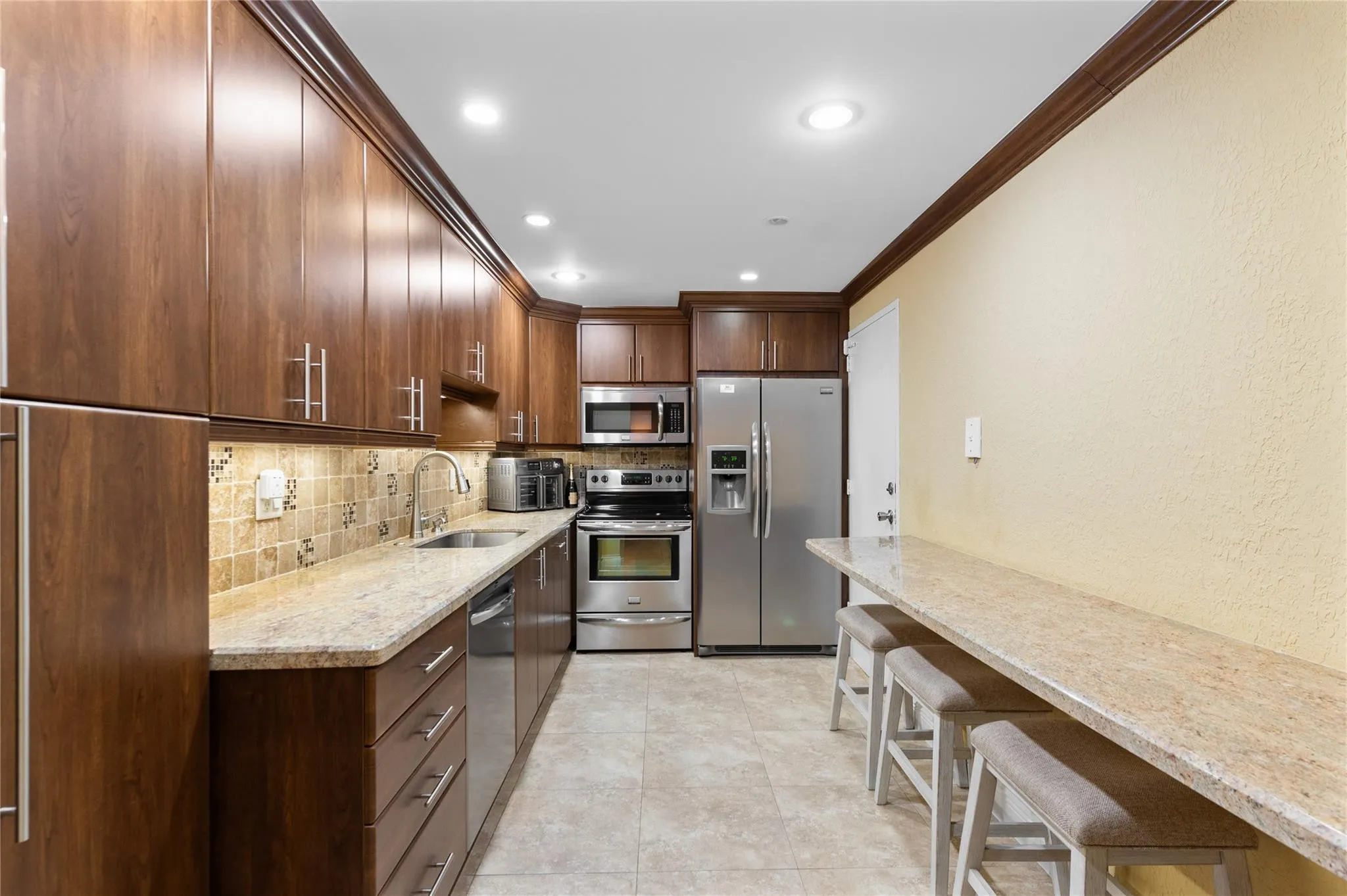 Galley kitchen featuring wood cabinetry, stainless steel appliances, recessed lighting, tile flooring, and light-toned countertops