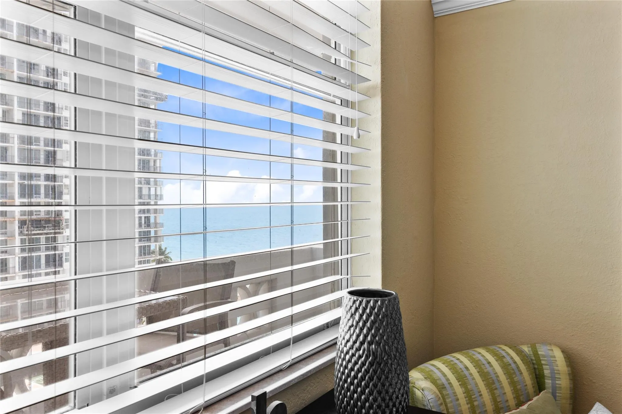 Expansive window with horizontal blinds, offering a glimpse of the ocean and adjacent high-rise buildings