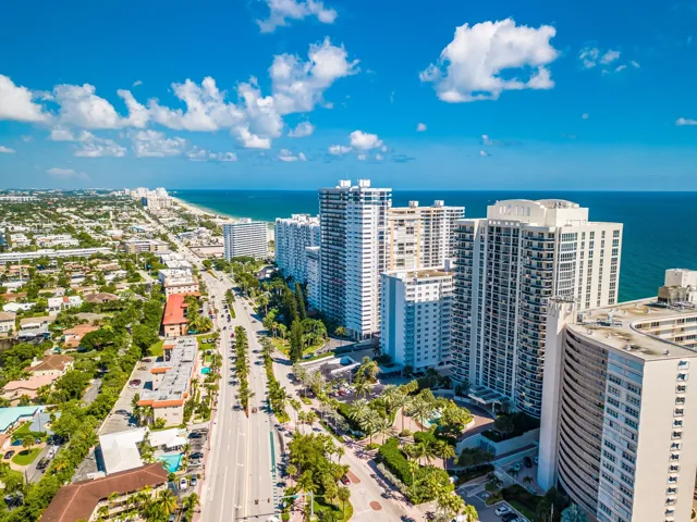 Coastal metropolitan setting featuring high-rise buildings, an expansive ocean, a broad sandy beach, and a tree-lined thoroughfare