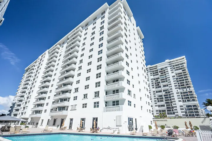 Multi-story building featuring numerous balconies with white railings, a clear blue swimming pool, and an adjacent building with similar architectural style