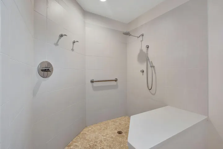 Tiled shower enclosure featuring a pebble-finish floor, a built-in shower bench, a handheld shower fixture, and a grab​​‌​​​​‌​​‌‌​‌‌​​​‌‌​‌​‌​‌​​​‌​​ bar