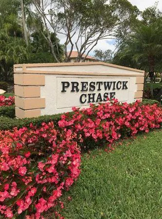 Community entrance featuring a stone sign with raised lettering, bordered by lush red flowering plants and verdant grass