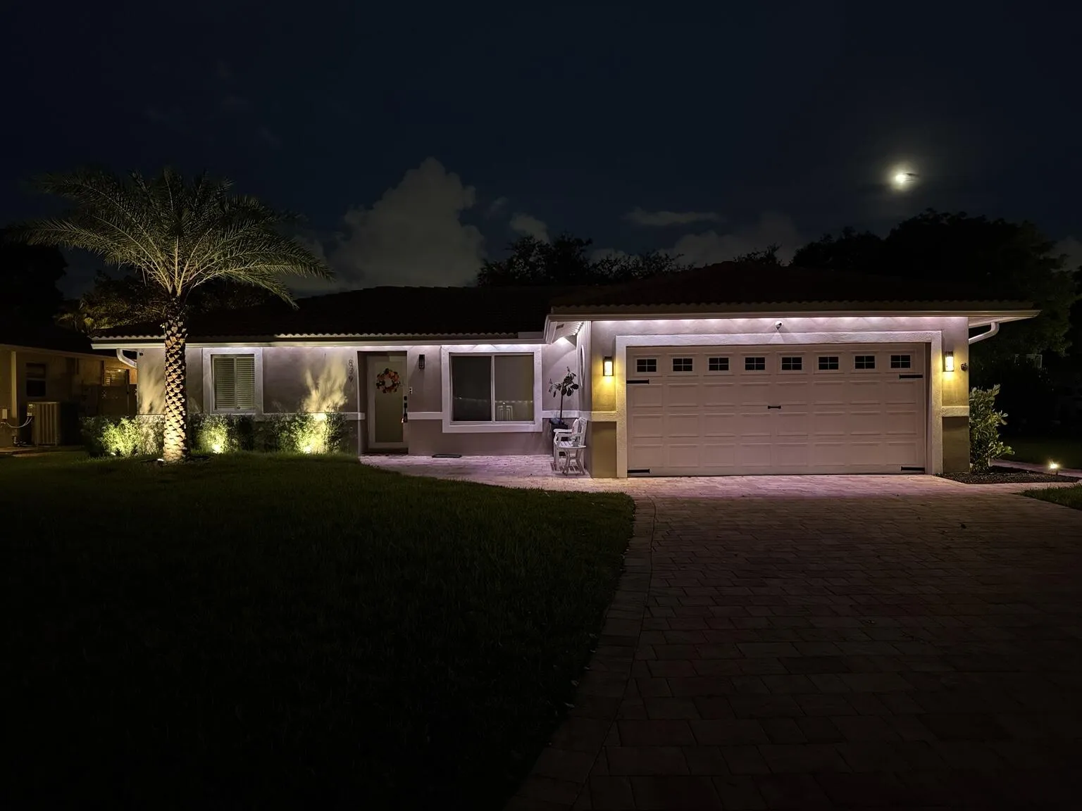 Modern single-story residence illuminated by professional landscape lighting, featuring a clean-lined architectural design with expansive glass panels, a two-car garage with paneled doors, brick paver driveway, mature palm tree specimen, and manicured lawn with perimeter hedging