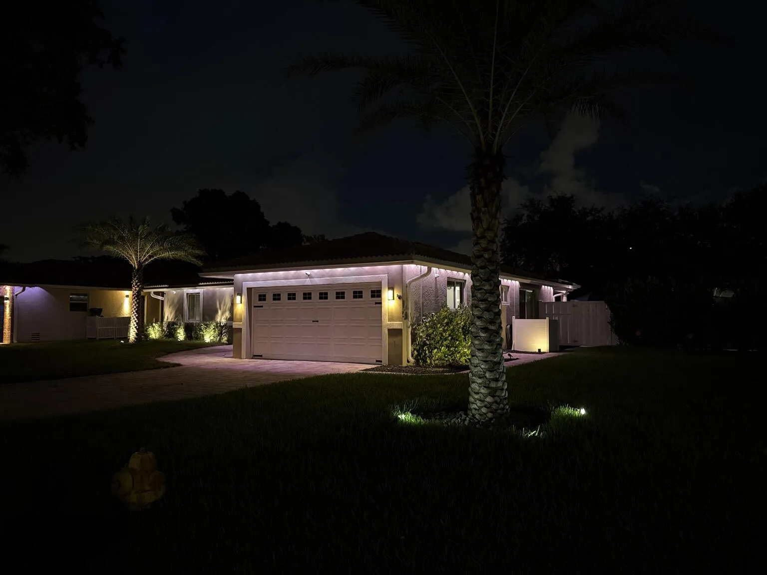 Recessed lighting, paver driveway, wall-mounted exterior fixtures, integrated garage door, and mature palm trees