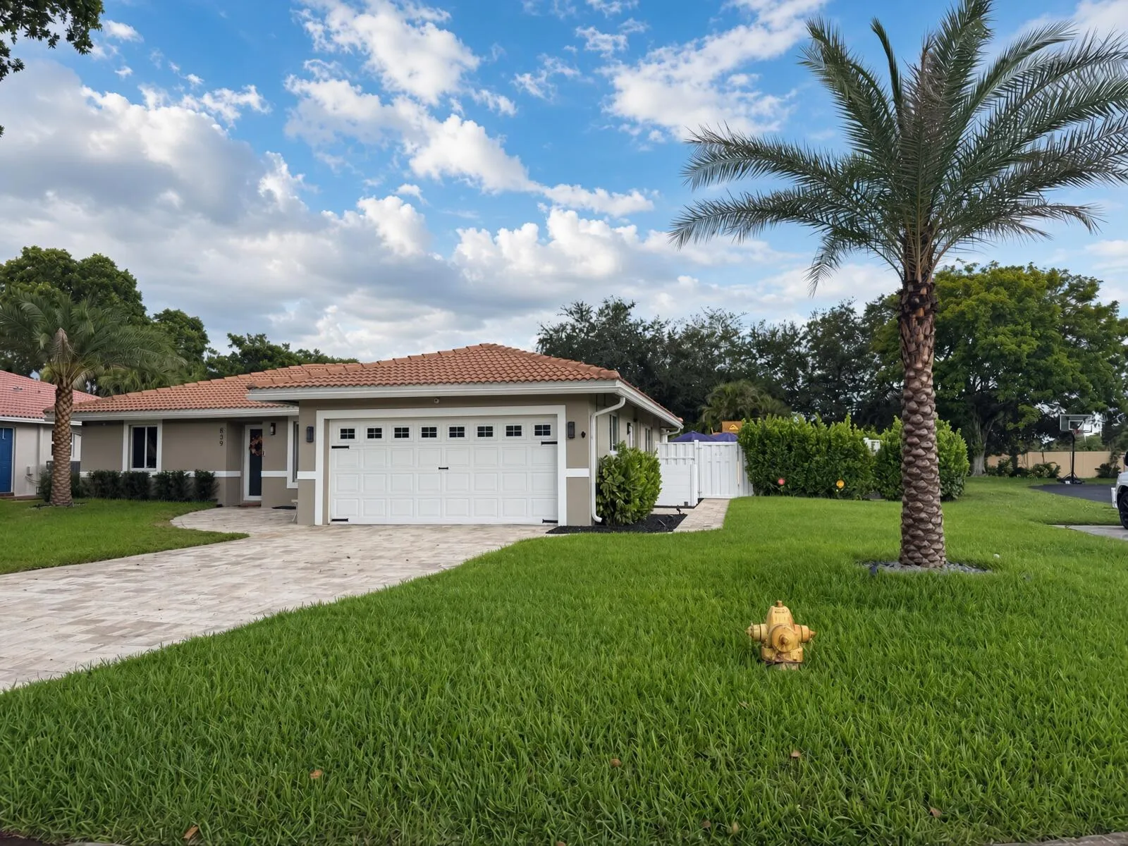 Terracotta tile roof, attached garage with white panel door, paver driveway, manicured lawn, and mature palm trees