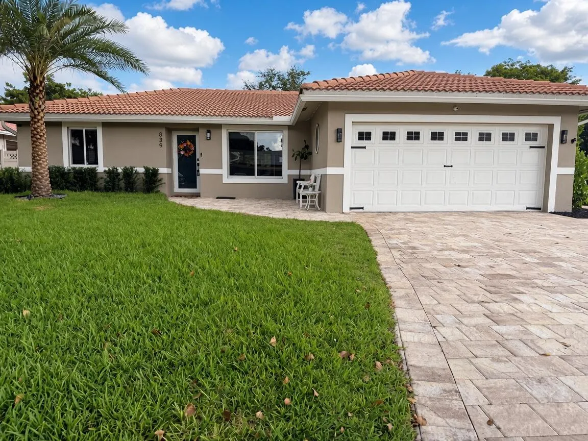 Welcoming single-story home featuring a spacious paver driveway, dual-bay garage with white panel doors, manicured lawn, and mature palm landscaping