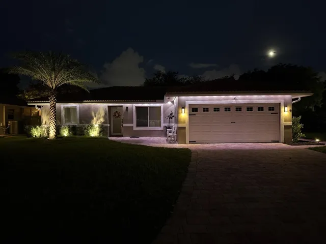 Modern single-story residence illuminated by professional landscape lighting, featuring a clean-lined architectural design with expansive glass panels, a two-car garage with paneled doors, brick paver driveway, mature palm tree specimen, and manicured lawn with perimeter hedging