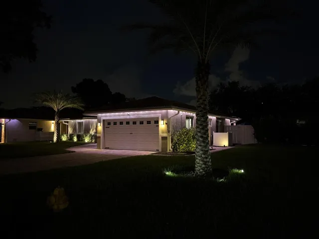 Recessed lighting, paver driveway, wall-mounted exterior fixtures, integrated garage door, and mature palm trees