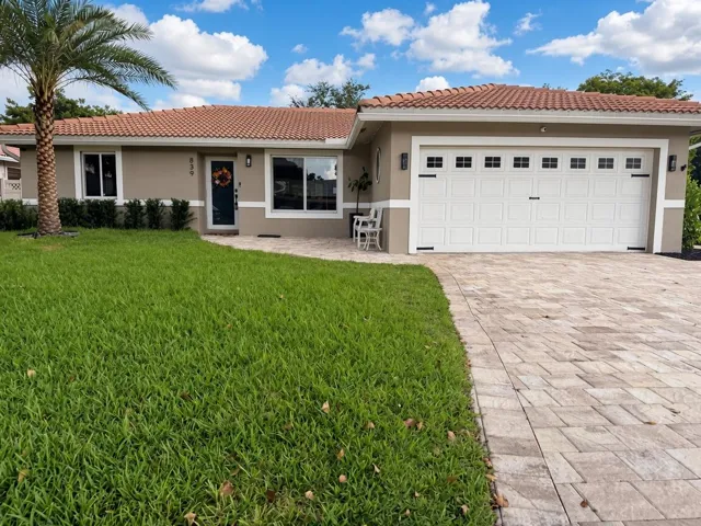 Welcoming single-story home featuring a spacious paver driveway, dual-bay garage with white panel doors, manicured lawn, and mature palm landscaping