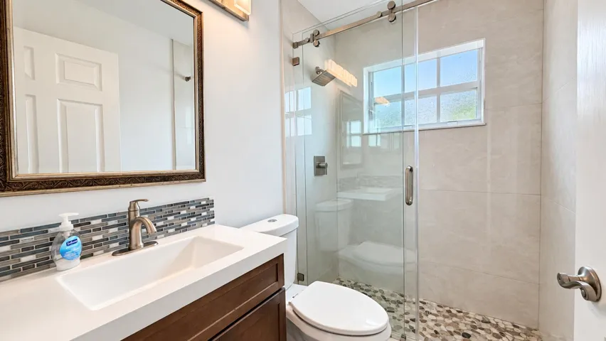Modern bathroom featuring a wood-finish vanity with an integrated sink, mosaic tile backsplash, and a frameless glass shower enclosure with a pebble floor