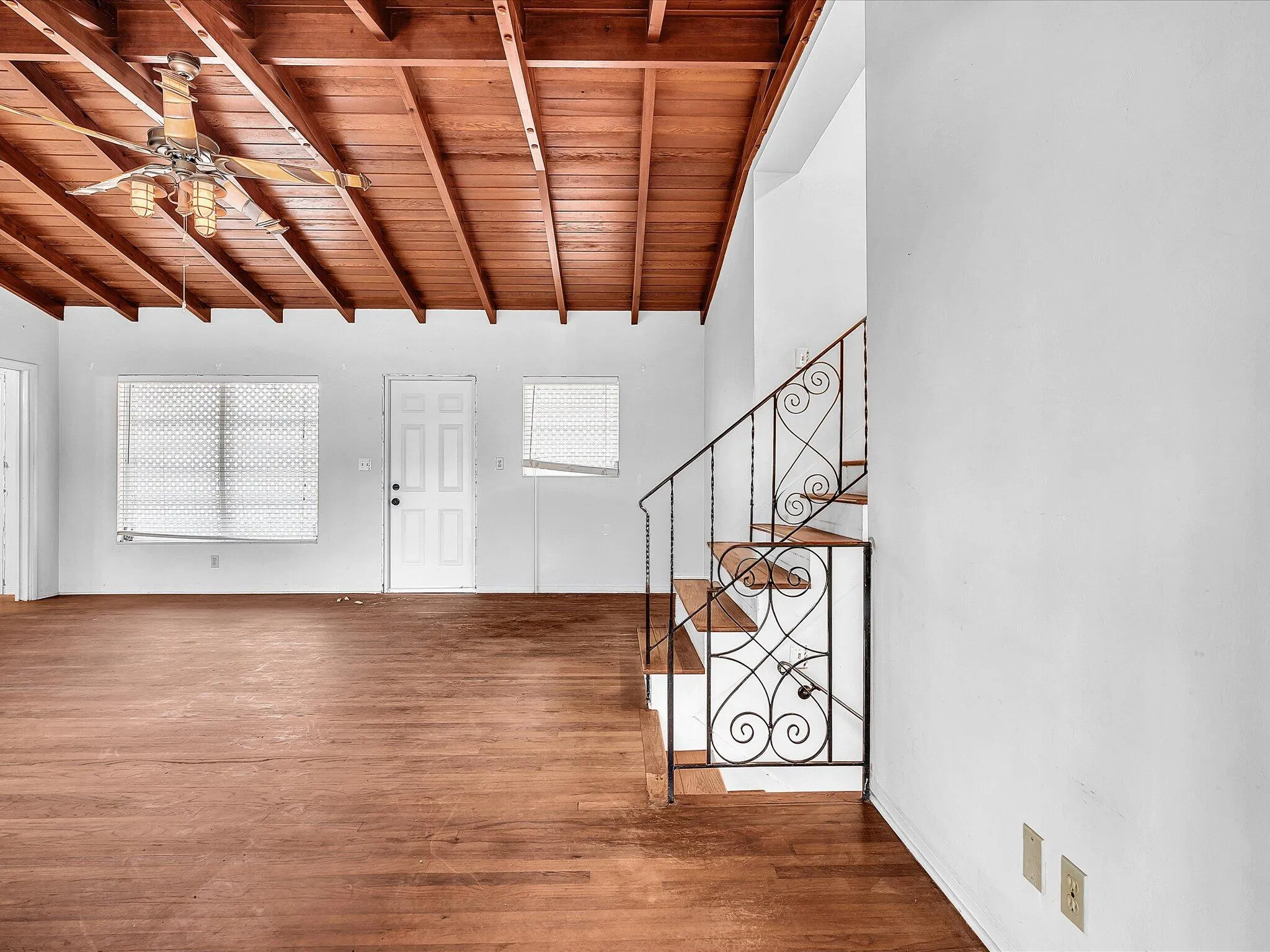 Exposed wood beam ceiling, wood-finish flooring, staircase with wrought iron railing, white paneled door, and ceiling fan with integrated lighting