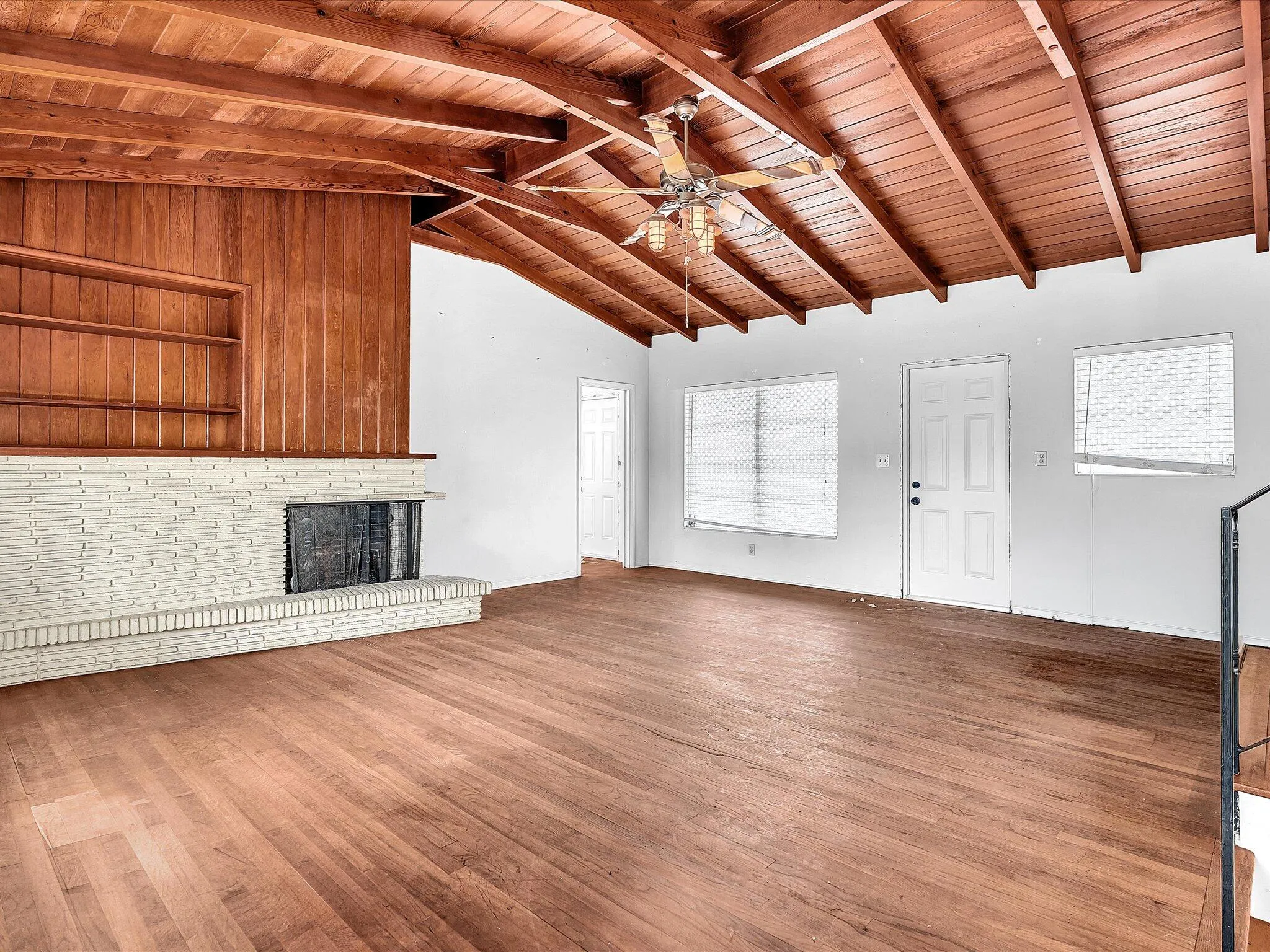 Vaulted wood-paneled ceiling with exposed beams, wood-finish flooring, white painted brick fireplace, wood-paneled accent wall with built-in shelving, and a ceiling fan
