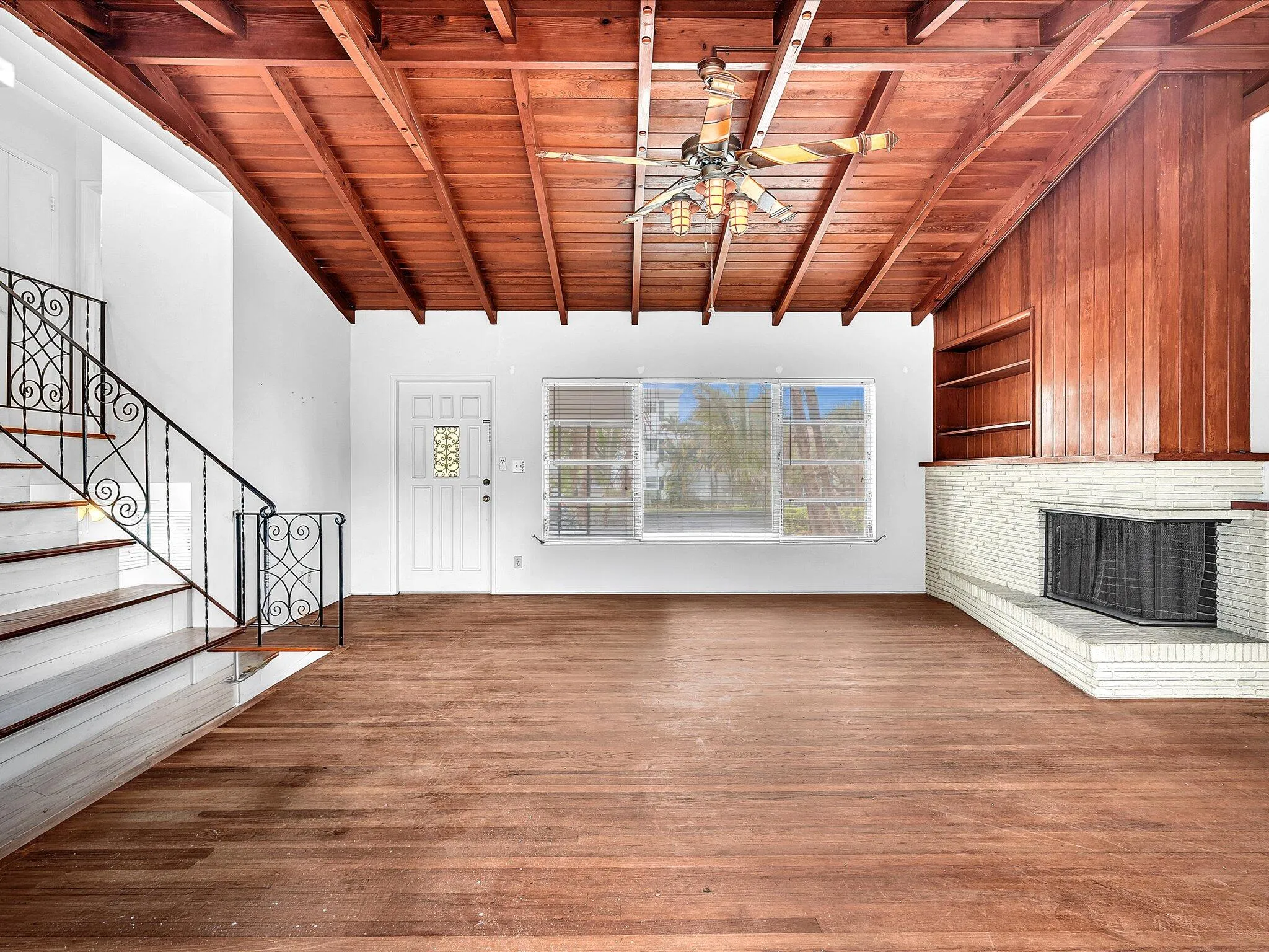 Exposed beam wood-finish ceiling, wood-finish flooring, white brick fireplace, built-in shelving, and an ornate wrought iron staircase
