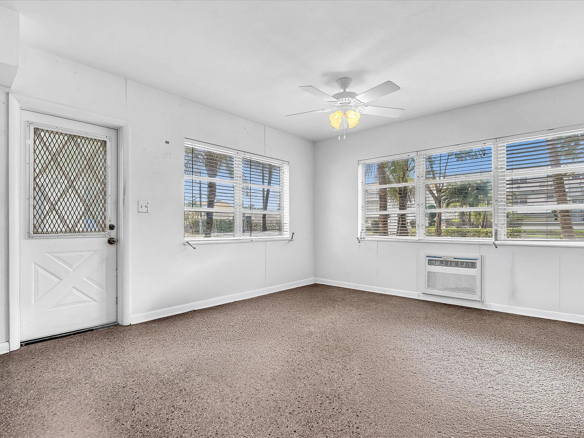 Multiple windows with horizontal blinds, ceiling fan with light fixture, wall-mounted air conditioning unit, and textured carpet flooring