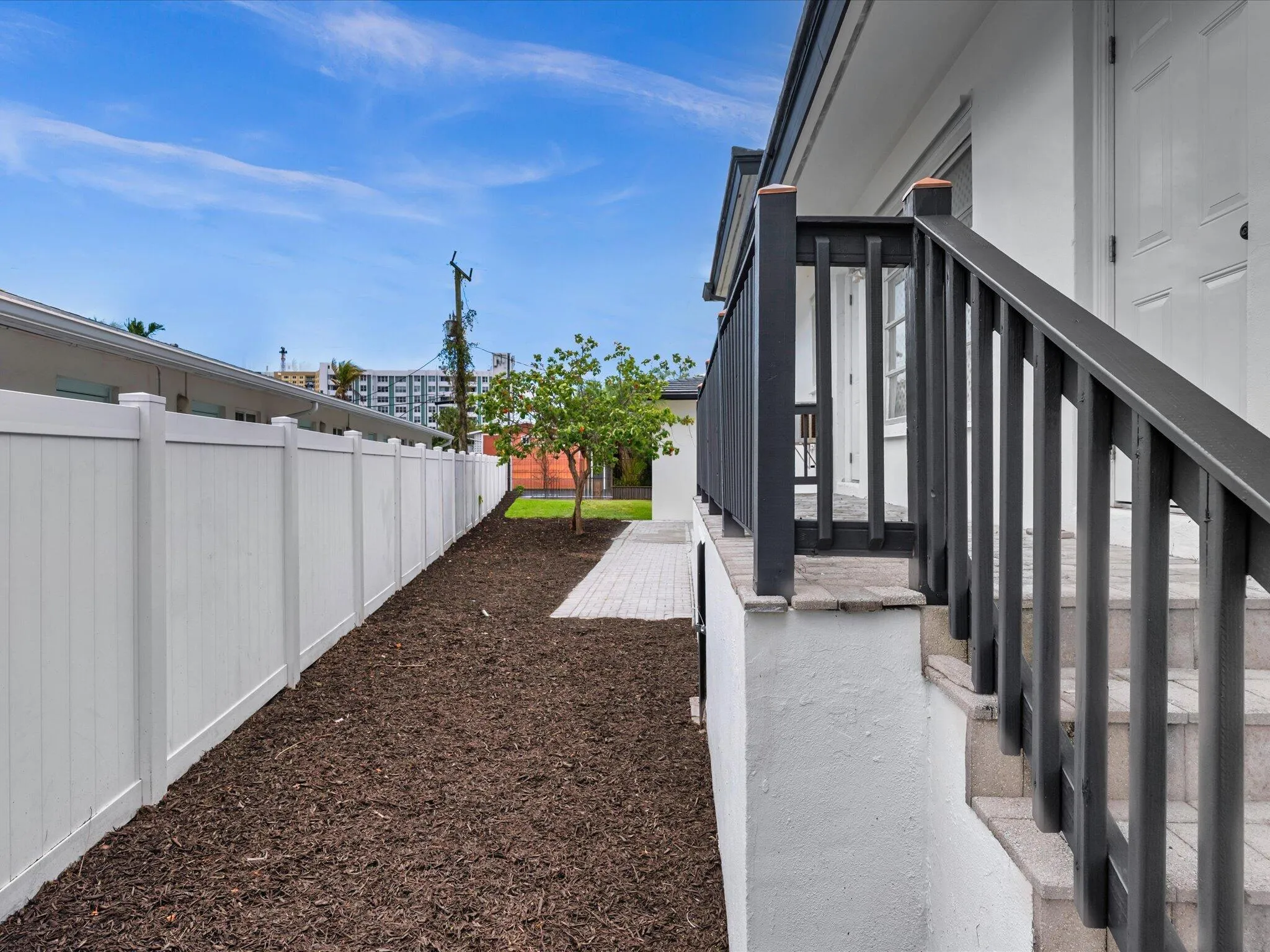 Exterior entryway with dark railings, stone steps, a white privacy fence, and a landscaped side yard