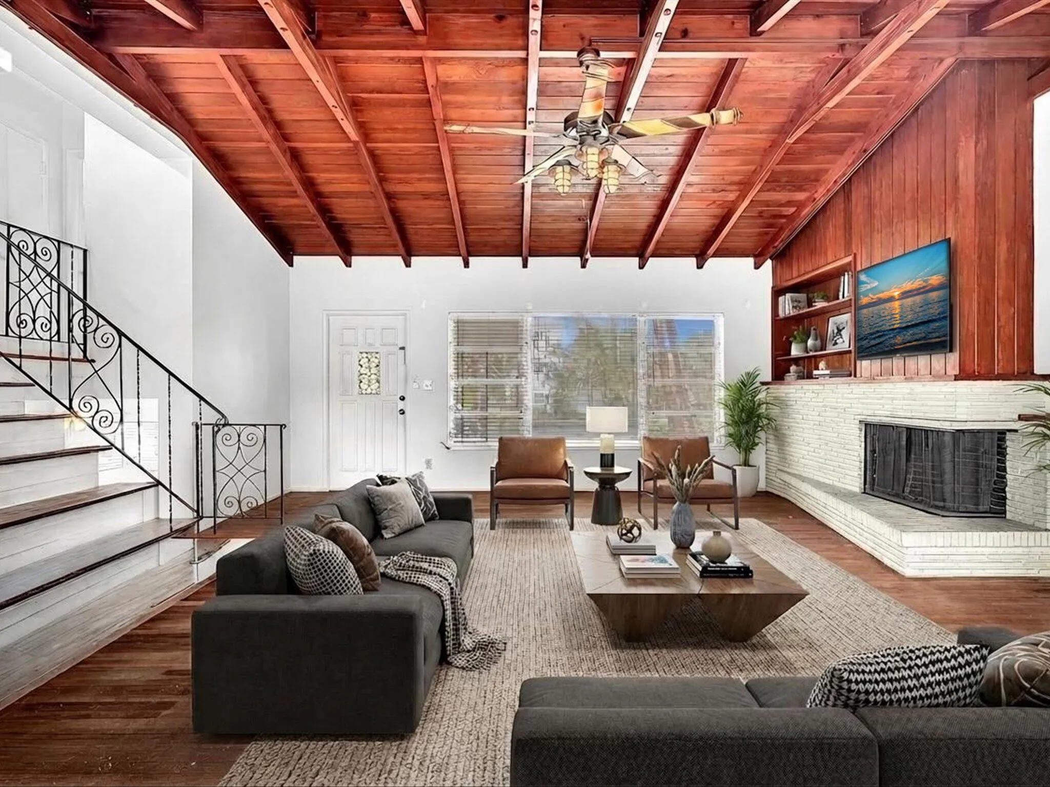 Vaulted wood-paneled ceiling with exposed beams, wood-finish flooring, white brick fireplace with built-in shelving, and an ornate wrought iron staircase