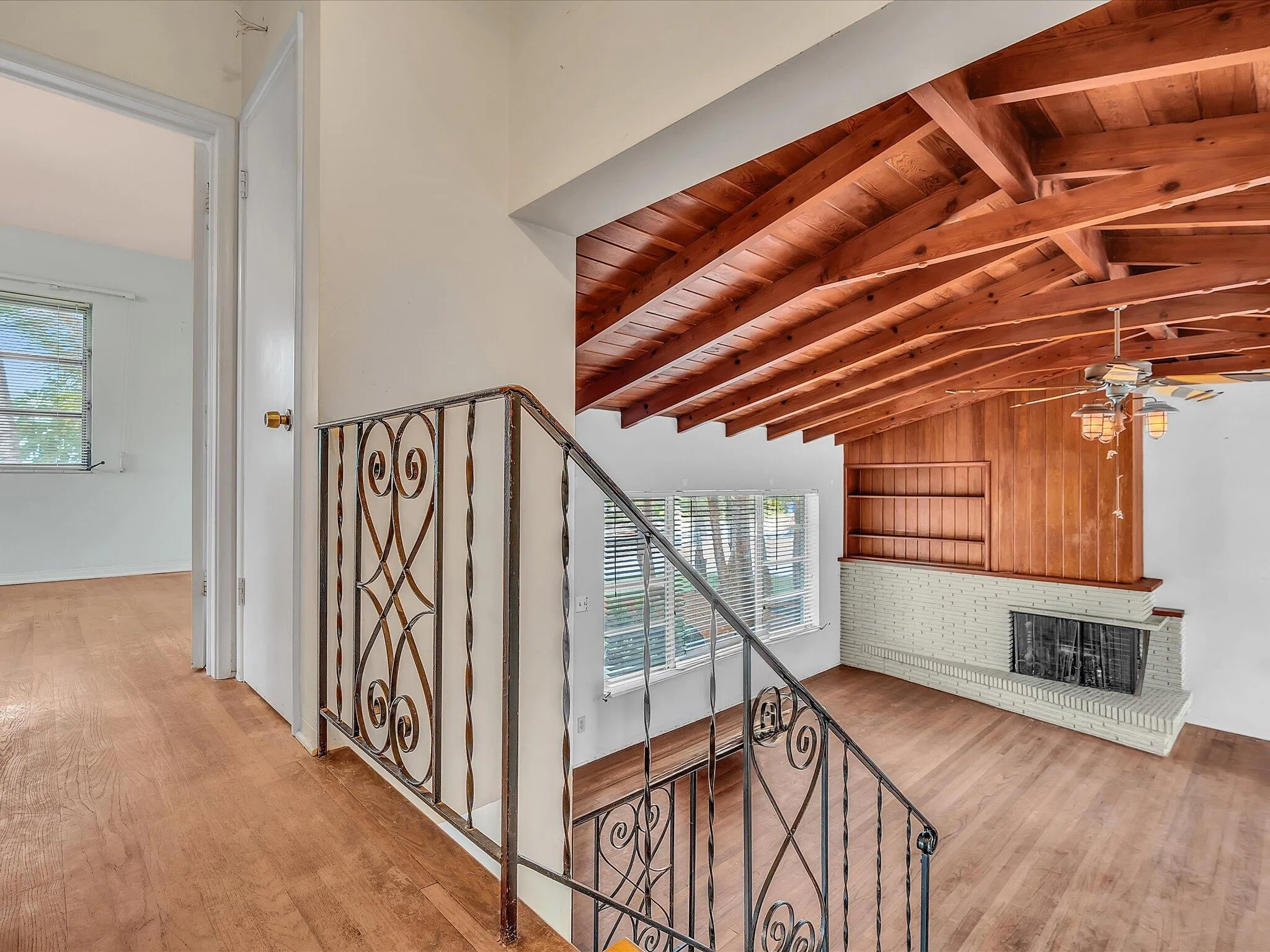 Vaulted wood-beam ceiling, wood-finish flooring, wrought iron staircase railing, built-in shelving, and a brick fireplace