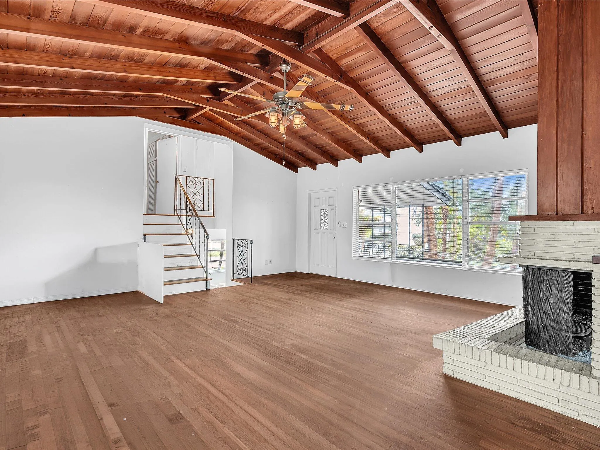 Vaulted wood-panel ceiling, wood-finish flooring, white brick fireplace, staircase with wrought iron railing, and a multi-pane window