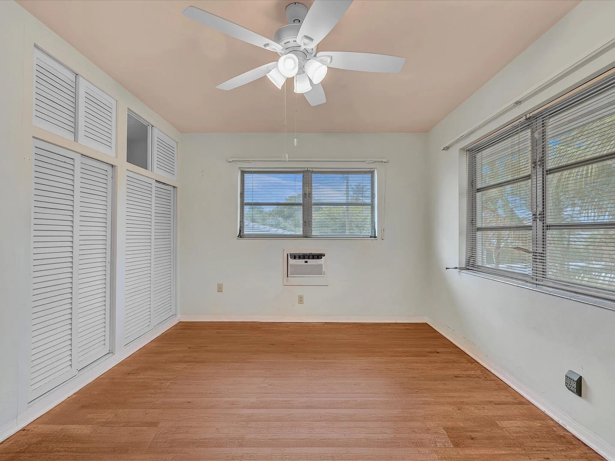 Wood-finish flooring, white louvered doors, ceiling fan with integrated lighting, multiple windows with blinds, and a built-in air conditioning unit