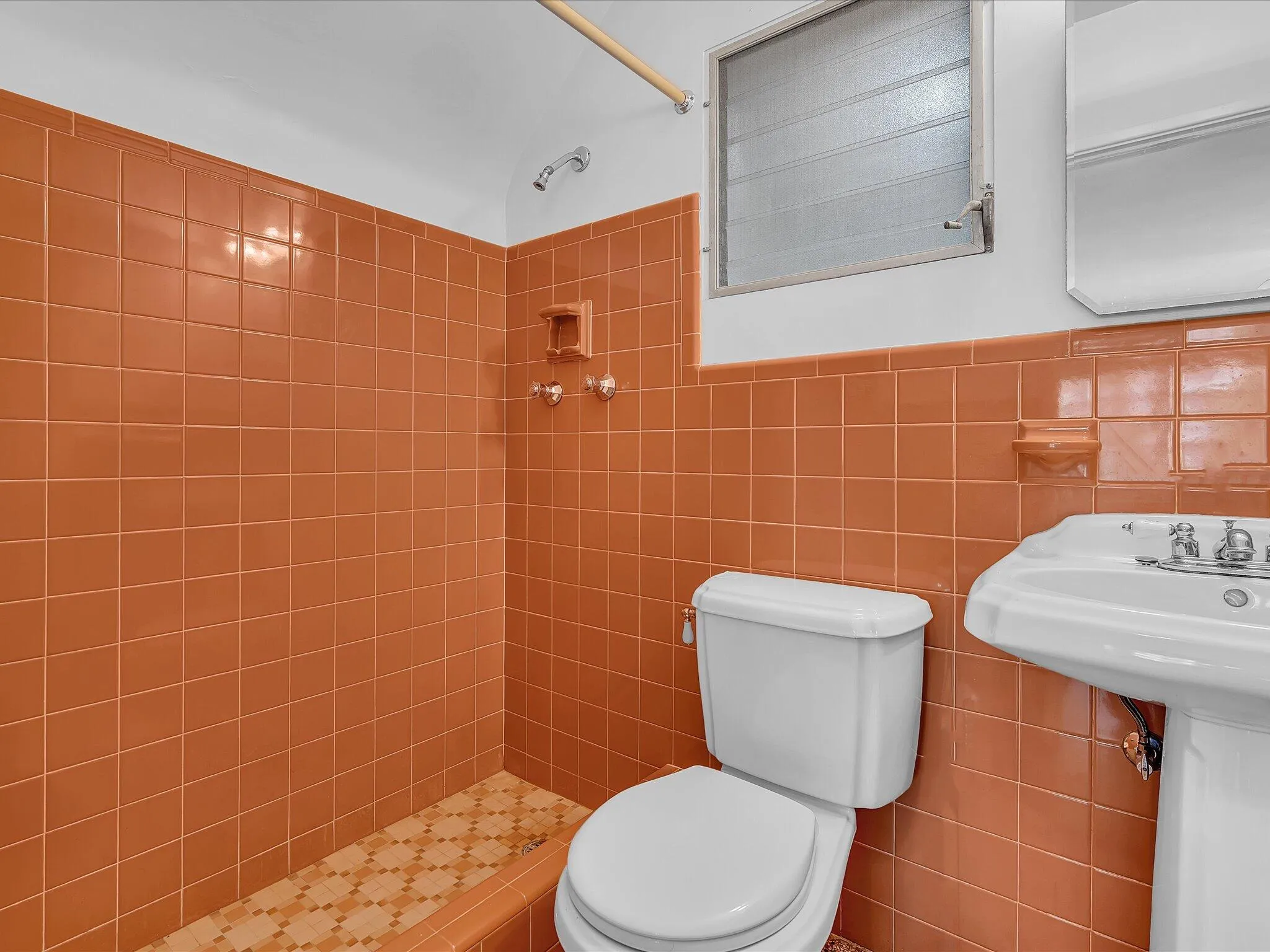 Terracotta-toned tiled walls, pedestal sink, shower with tiled floor, and a louvered window