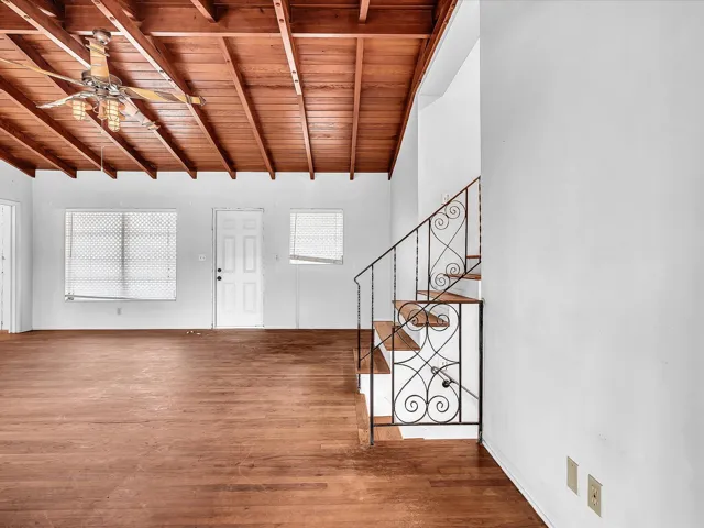 Exposed wood beam ceiling, wood-finish flooring, staircase with wrought iron railing, white paneled door, and ceiling fan with integrated lighting