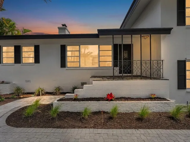 White exterior with dark shutters, covered entry with decorative wrought iron railing, paved walkway, and landscaped garden beds