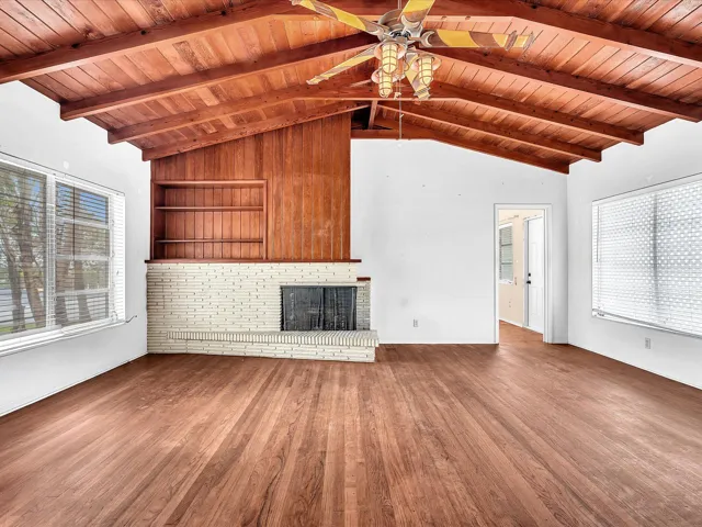 Vaulted wood-paneled ceiling, wood-finish flooring, white brick fireplace, built-in shelving, and large windows