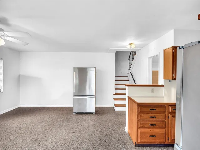 Stainless steel refrigerator, wood cabinetry with dark hardware, light countertops, staircase with matching wood treads and black handrail, and a ceiling fan