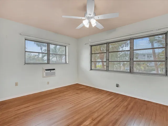 Hardwood flooring, multiple windows providing natural light, a ceiling fan with integrated lighting, and a wall-mounted air conditioning unit