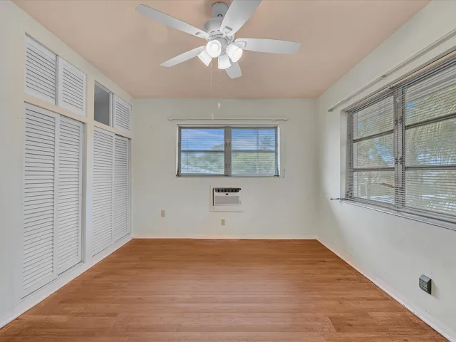 Wood-finish flooring, white louvered doors, ceiling fan with integrated lighting, multiple windows with blinds, and a built-in air conditioning unit