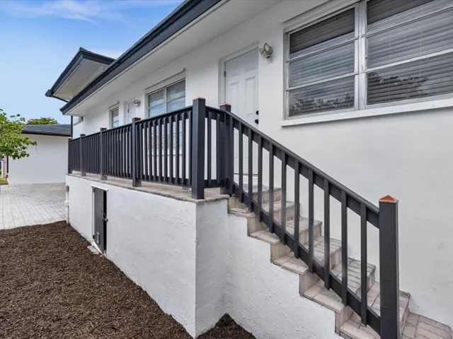 White stucco exterior with a dark roofline, featuring exterior stairs with dark railings, a paved walkway, and a small built-in access door