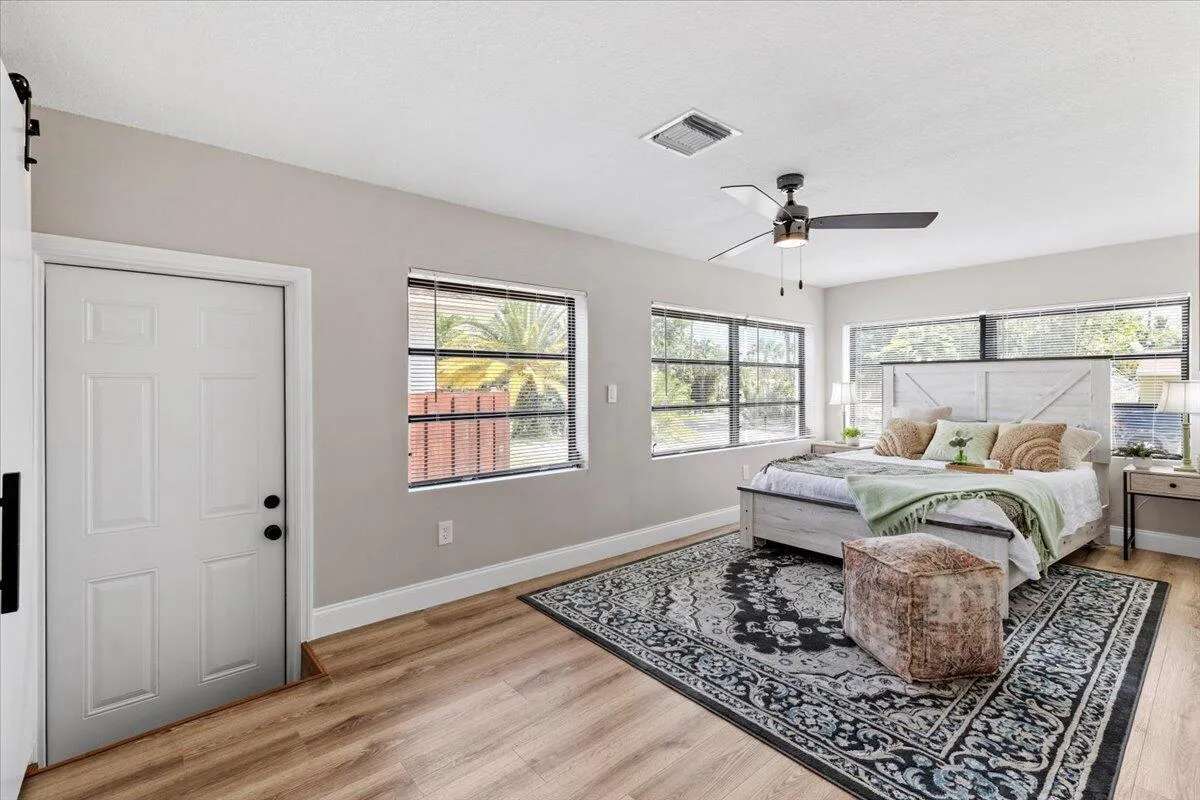 Wood-finish flooring throughout, a ceiling fan with integrated lighting, multiple windows providing natural illumination, and a white paneled door with black hardware