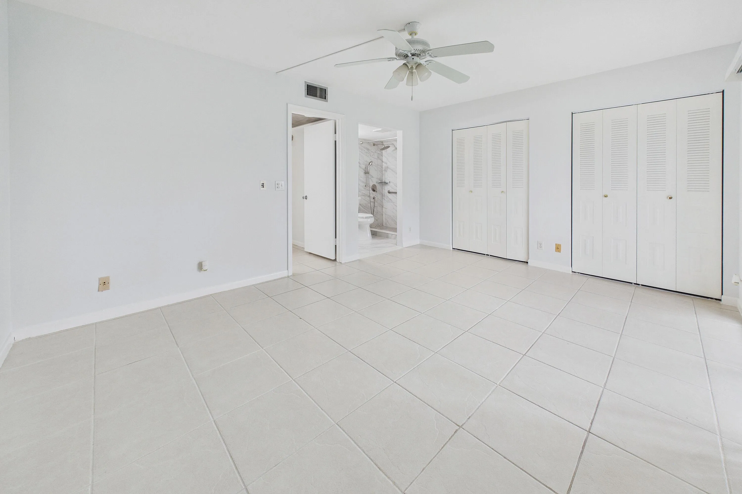 Light-toned tile flooring, two louvered bifold closet doors, a ceiling fan, and a neutral wall color