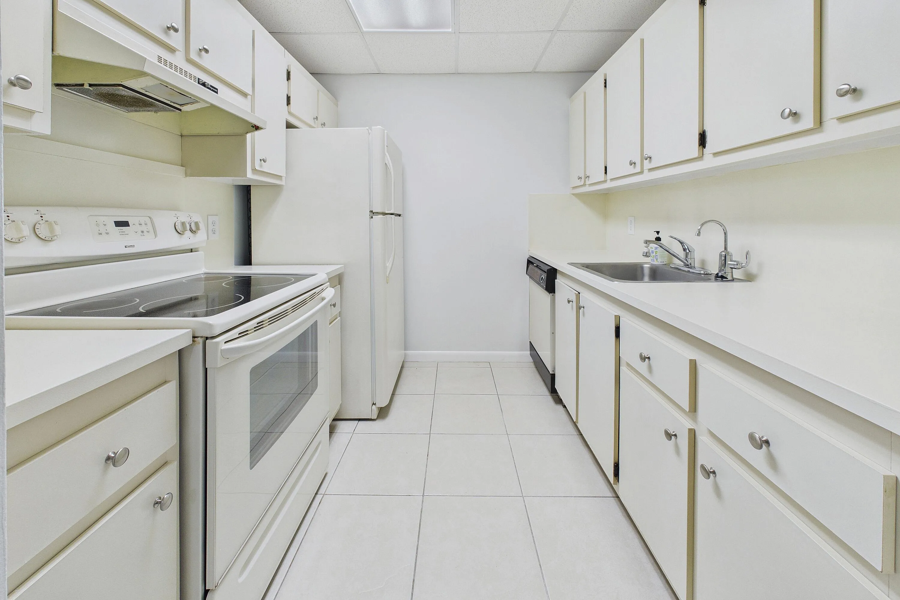 Galley kitchen featuring white cabinetry, solid surface countertops, tile flooring, a stainless steel sink, and white appliances