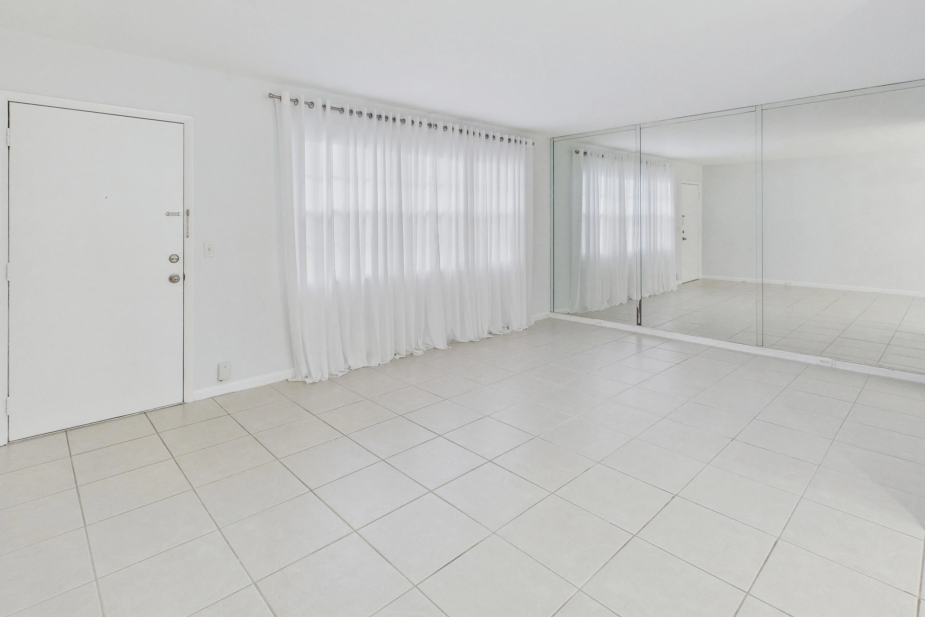 White tile flooring, full-height mirrored wall, and a window with sheer white curtains.