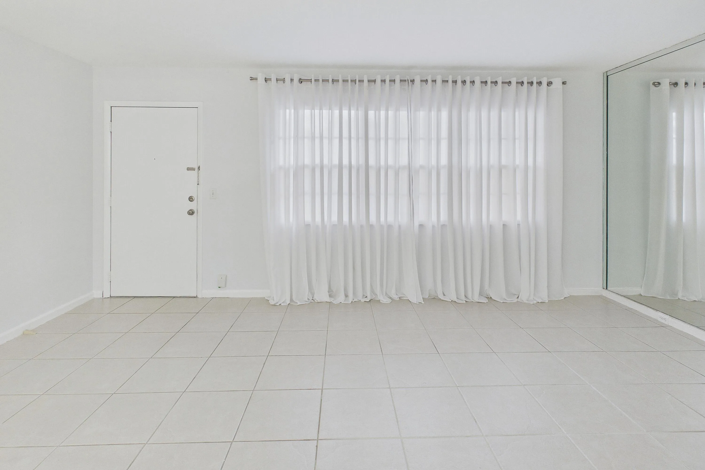Tile flooring, white walls, full-length mirror, window with sheer curtains, solid entry door