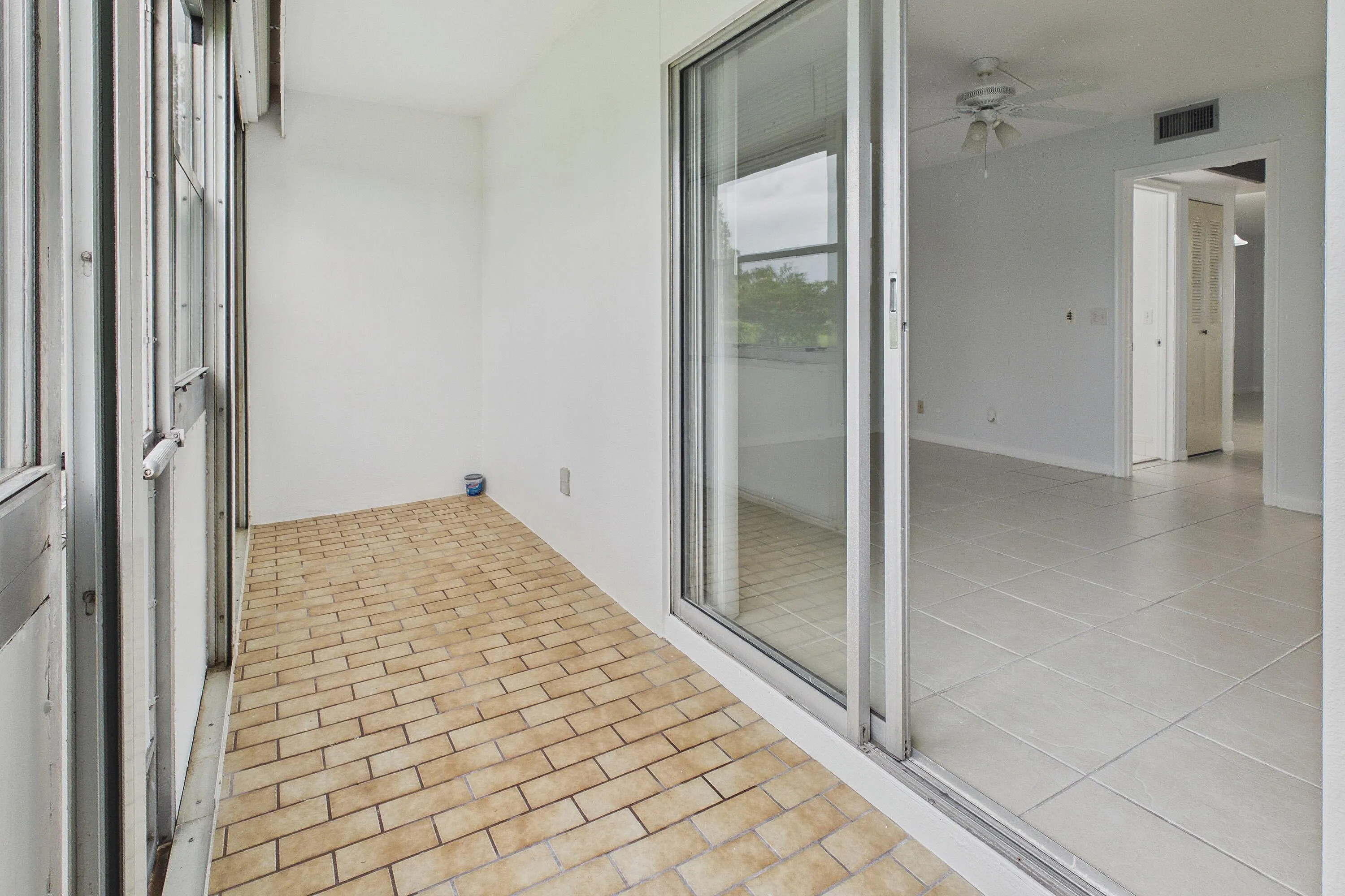 Enclosed and covered patio with brick-pattern flooring and white walls, offering access to an interior space with light-toned tile flooring and a ceiling fan