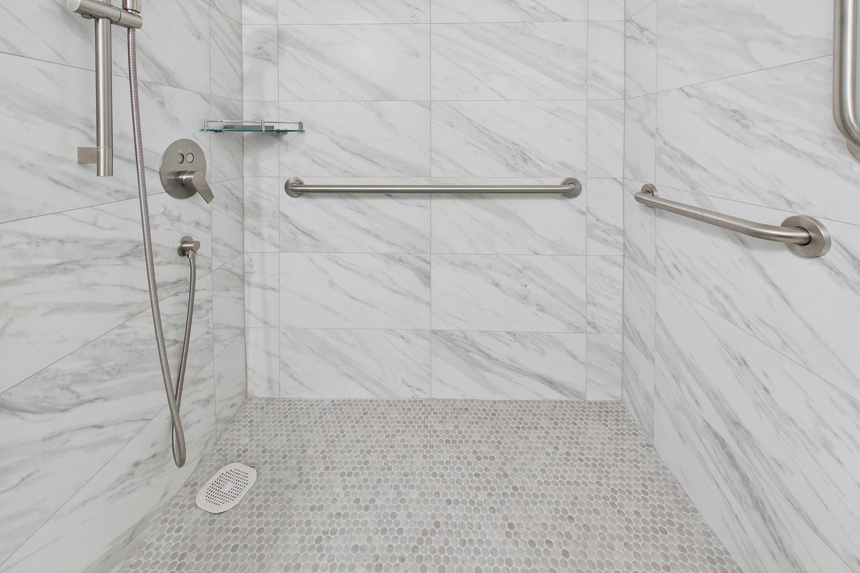 Renovated handicap accessible white marble-look wall tile, pebble-finish shower floor, brushed nickel shower fixtures, glass shower shelf, and integrated grab bars