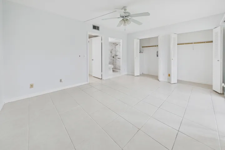 White tile flooring, dual bi-fold closet doors, ceiling fan with light fixture, and a white interior door leading to a bathroom with a tiled shower