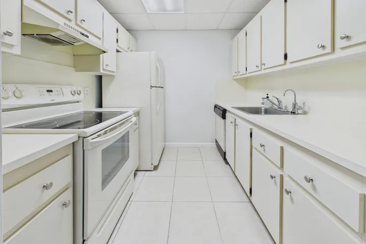 Galley kitchen featuring white cabinetry, solid surface countertops, tile flooring, a stainless steel sink, and white appliances
