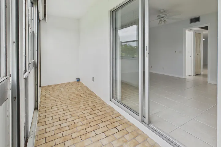 Enclosed and covered patio with brick-pattern flooring and white walls, offering access to an interior space with light-toned tile flooring and a ceiling fan