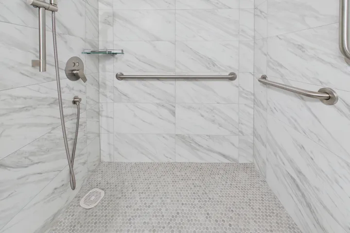 Renovated handicap accessible white marble-look wall tile, pebble-finish shower floor, brushed nickel shower fixtures, glass shower shelf, and integrated grab bars