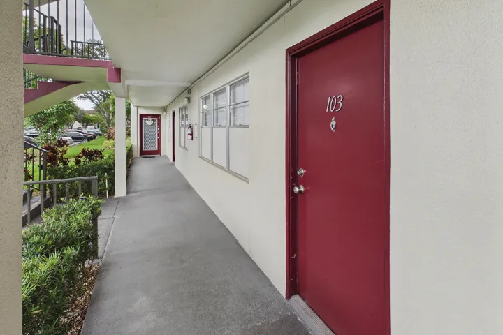 Covered exterior walkway featuring a solid entry door with contrasting trim, multiple windows, and exterior foliage