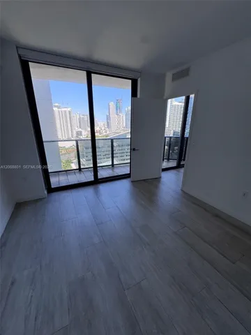 Floor-to-ceiling glass sliding doors, private balcony, wood-finish flooring, and urban skyline backdrop