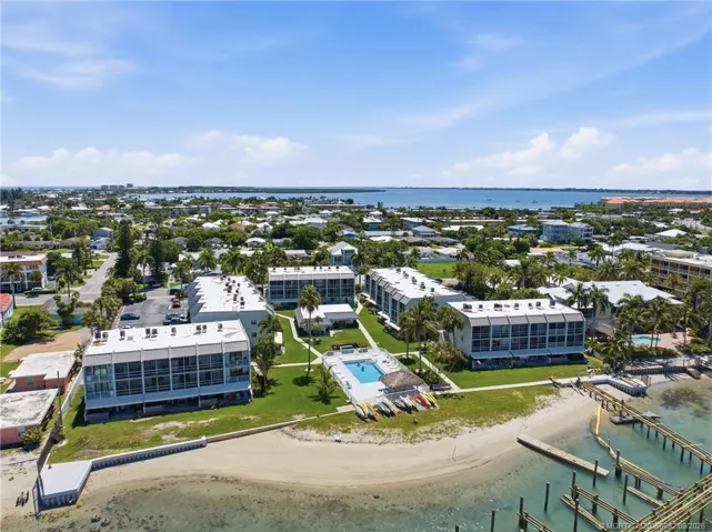 Aerial view showcasing the community’s waterfront setting, pool, sandy shoreline, and surrounding coastal neighborhood.
